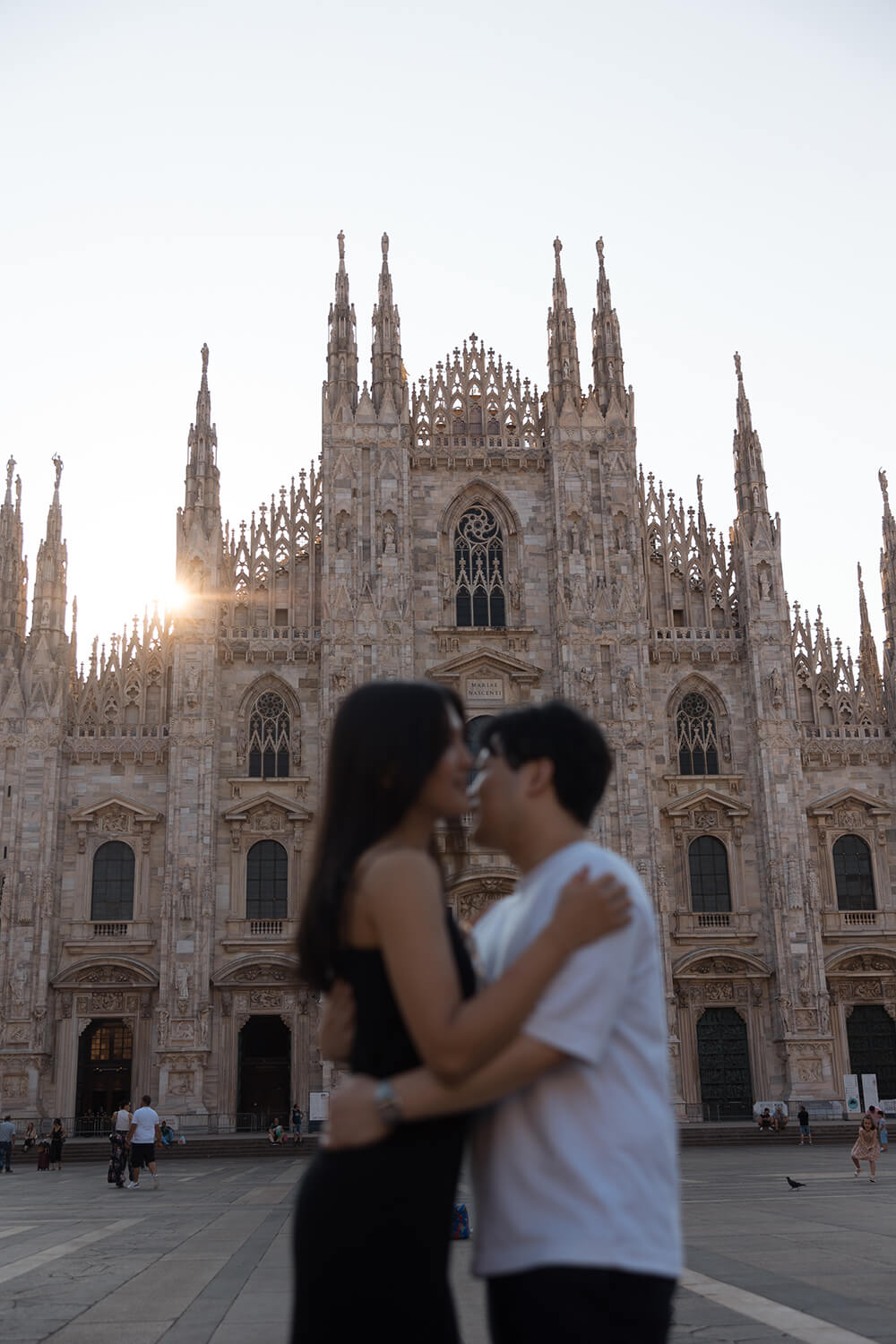 イタリア・ミラノのドゥオーモ広場で撮影された、映画のワンシーンのようなカップルフォト。朝日が差し込む美しいドゥオーモにピントを合わせ、手前で抱き合い見つめ合うふたりをドラマチックに切り取った、Totte（トッテ）のヨーロッパ私服ロケーション撮影。