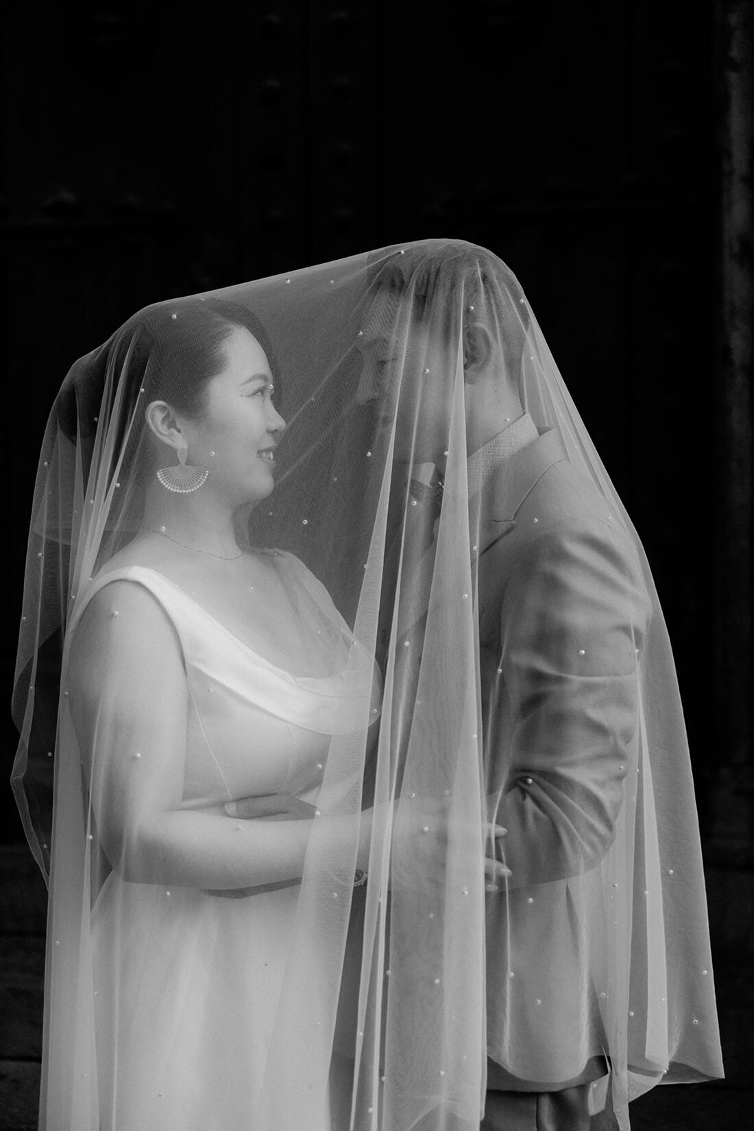 バルセロナ旧市街の静かな片隅で、ベールの下で見つめ合う新郎新婦のロマンチックなモノクロ写真。お二人だけの特別な時間を切り取った、タイムレスでエモーショナルなウェディングフォト。歴史ある街並みに溶け込む、芸術的で温かな前撮りカット。