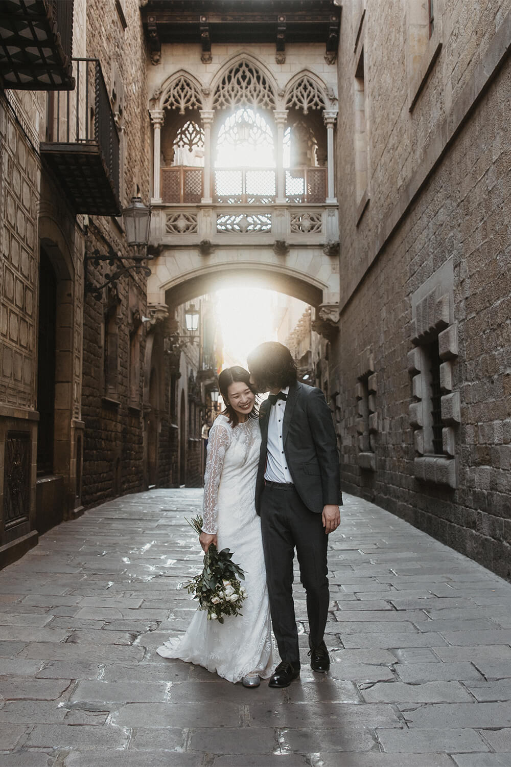 バルセロナ旧市街の象徴、ビスベ橋の下で光に包まれる新郎新婦。中世の面影を残すゴシック地区の美しいアーチから差し込む光を活かした、ロマンチックなウェディングフォト。Totteがプロデュースする、映画のワンシーンのようなバルセロナ前撮りの一枚。