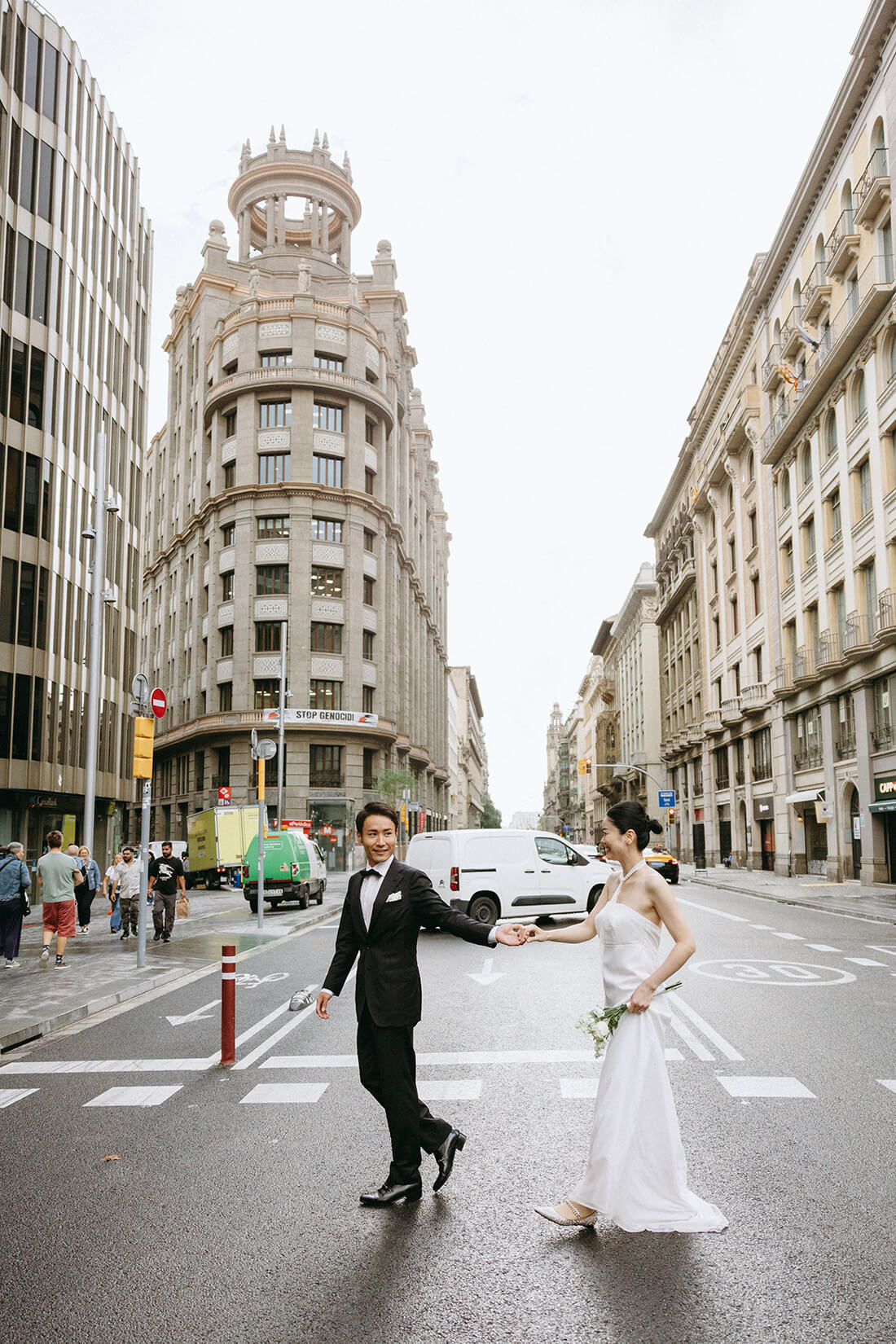 バルセロナの街角で、横断歩道を渡る新郎新婦のスタイリッシュなウェディングフォト。活気ある街の日常に溶け込みながら、おふたりの幸せな歩みを捉えたロケーション撮影。スペイン旅行の空気感をそのままに、軽やかで映画的な前撮りシーン。