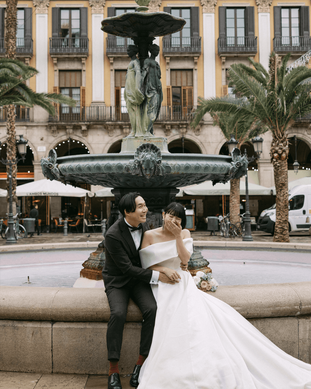 スペイン・バルセロナの旧市街にあるレイアール広場の美しい噴水に座り、楽しそうに笑い合う新郎新婦のウェディングフォト。クラシカルな歴史的建造物とパームツリーに囲まれた、ヨーロッパでの海外前撮り・ロケーション撮影（Totte）。