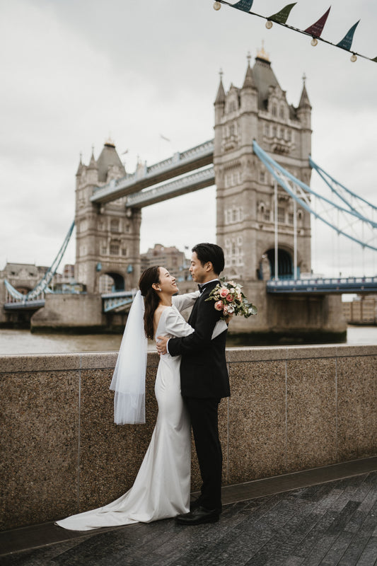 ロンドンの象徴的なランドマーク、タワー・ブリッジ（Tower Bridge）を雄大な背景に、ウェディングブーケを手に持ち、愛おしそうに見つめ合う新郎新婦。テムズ川のほとりで撮影された、ロイヤルでクラシックな雰囲気漂う、ELLE研修修了プランナー監修・Totteのドラマチックなフォトウェディング・前撮り写真。