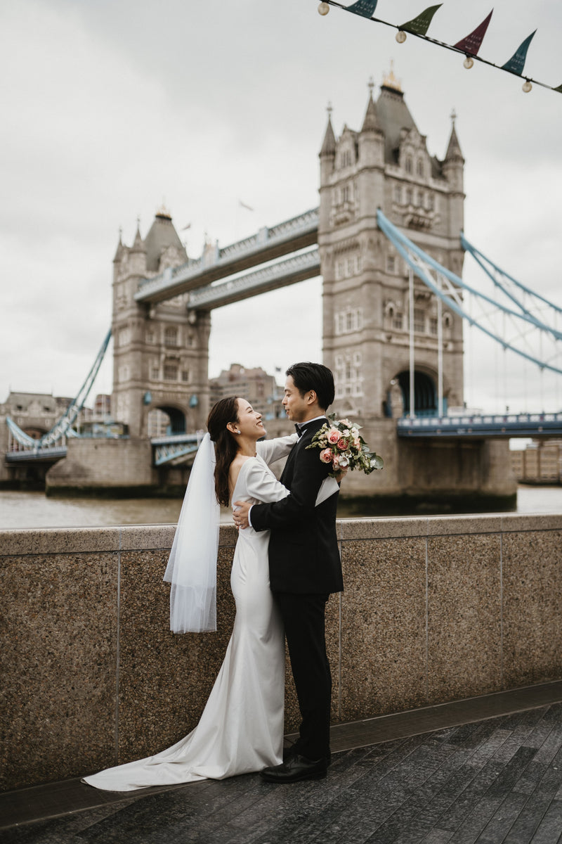 ロンドンの象徴的なランドマーク、タワー・ブリッジ（Tower Bridge）を雄大な背景に、ウェディングブーケを手に持ち、愛おしそうに見つめ合う新郎新婦。テムズ川のほとりで撮影された、ロイヤルでクラシックな雰囲気漂う、ELLE研修修了プランナー監修・Totteのドラマチックなフォトウェディング・前撮り写真。