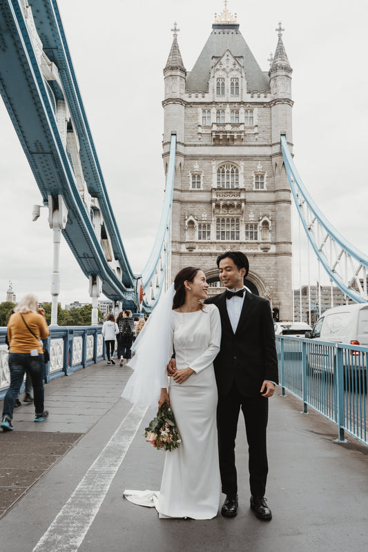 ロンドン・タワーブリッジ（Tower Bridge）の歩道にて、ヴィクトリア・ゴシック様式の壮麗なタワー（主塔）を背景に佇む新郎新婦。行き交う人々やロンドンの日常風景が溶け込んだ、映画のワンシーンのように自然体でドラマチックなフォトウェディング・前撮り写真。ELLE研修修了プランナー監修・Totte撮影。