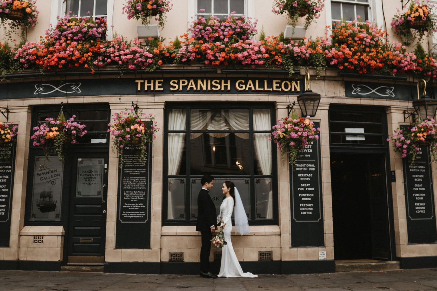 ロンドンの街角にある伝統的なパブ（Pub）の前で、外壁を彩る華やかな暖色系のフラワーデコレーションを背景に佇む新郎新婦。イギリスらしい重厚な木造建築と、鮮やかな花々が織りなすフォトジェニックなロケーションで撮影された、ELLE研修修了プランナー監修・Totteのおしゃれで英国情緒あふれるフォトウェディング・前撮り写真。