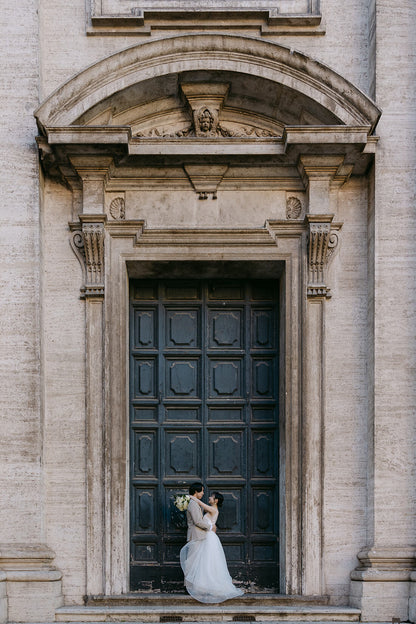イタリア・ローマの歴史的な建物の重厚で美しい木製ドアの前に佇む新郎新婦。古代の街並みに溶け込むアンティークな扉の質感と、洗練されたおふたりの姿を対比させた、ELLE研修修了プランナー監修・Totteの映画のようなフォトウェディング・ロケーション撮影。