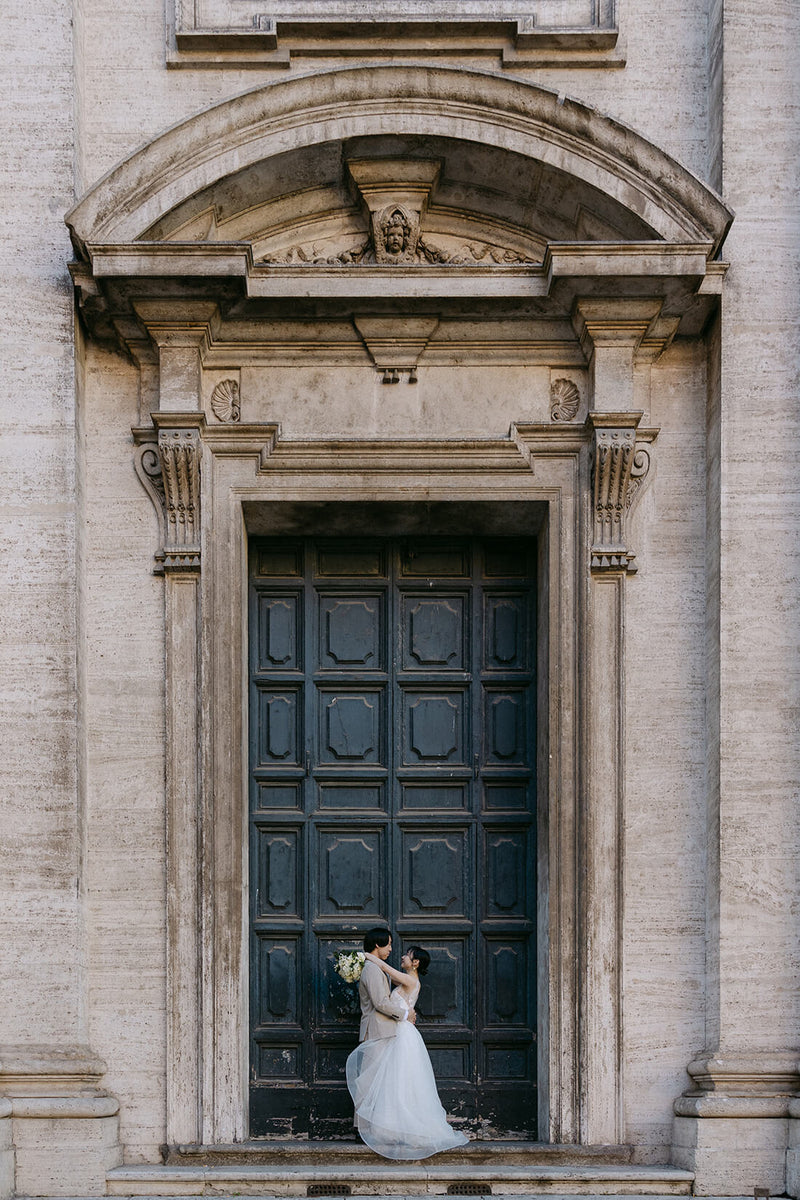 イタリア・ローマの歴史的な建物の重厚で美しい木製ドアの前に佇む新郎新婦。古代の街並みに溶け込むアンティークな扉の質感と、洗練されたおふたりの姿を対比させた、ELLE研修修了プランナー監修・Totteの映画のようなフォトウェディング・ロケーション撮影。