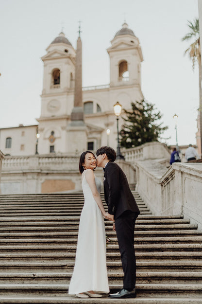 ローマの象徴・スペイン広場（Piazza di Spagna）の大階段にて、手を取り合い、新郎が新婦の頬に優しくキスをするロマンチックな一枚。映画『ローマの休日』の舞台となった憧れのロケーションで、ELLE研修修了プランナー監修・Totteが叶える、ドラマチックで幸せあふれるフォトウェディング・前撮り。