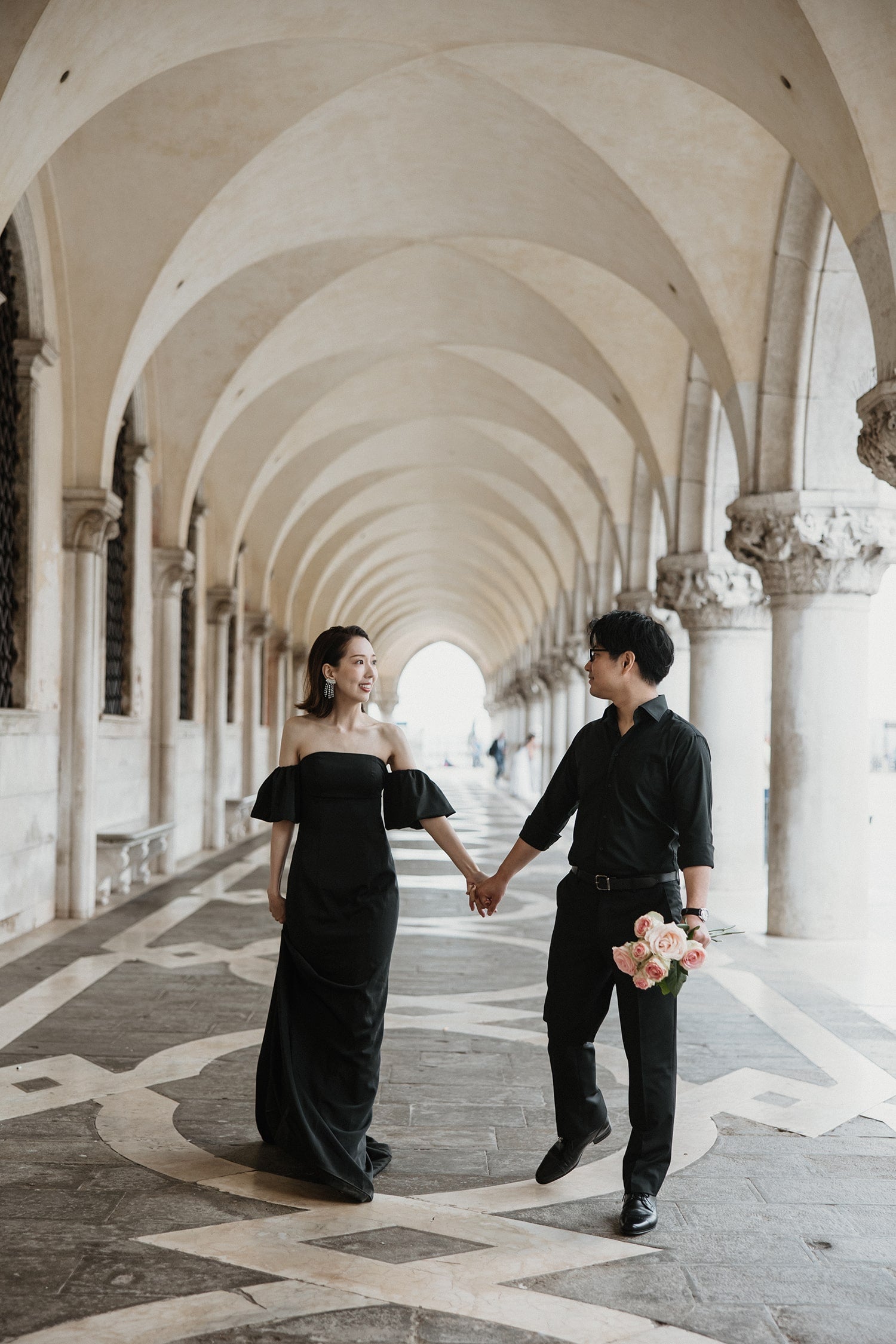 イタリア・ベネチアのドゥカーレ宮殿（Palazzo Ducale）の回廊にて、連続する美しいアーチの下を手をつなぎ優雅に歩く新郎新婦。ゴシック様式の建築美と光のコントラストが印象的な、ELLE研修修了プランナー監修・Totteの洗練されたフォトウェディング・前撮り写真。