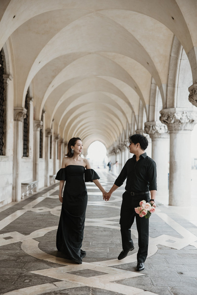 イタリア・ベネチアのドゥカーレ宮殿（Palazzo Ducale）の回廊にて、連続する美しいアーチの下を手をつなぎ優雅に歩く新郎新婦。ゴシック様式の建築美と光のコントラストが印象的な、ELLE研修修了プランナー監修・Totteの洗練されたフォトウェディング・前撮り写真。